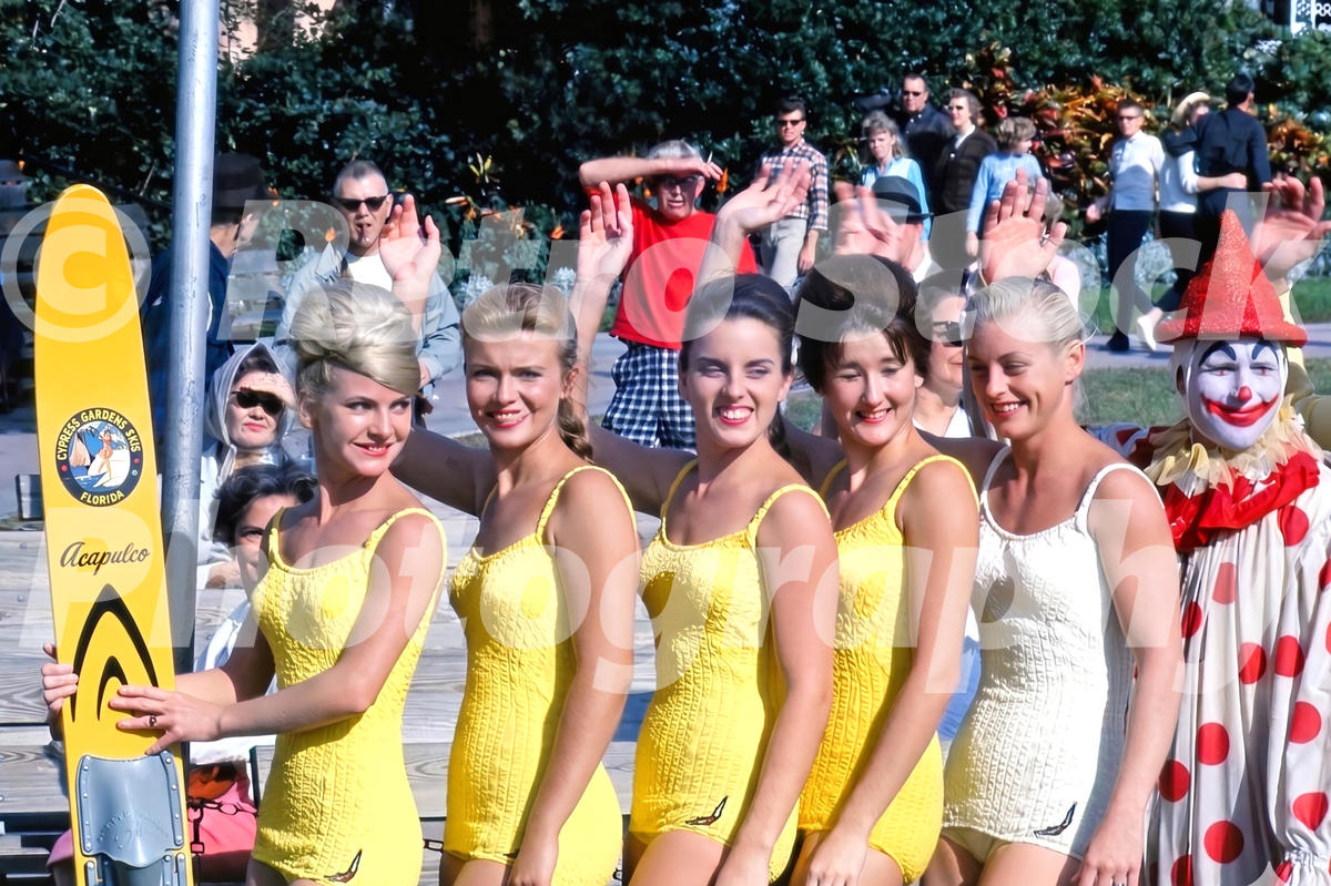 A 1960s color photo of five female water skiers at Cypress Gardens, Florida, wearing yellow and white textured swimsuits, posing with a yellow "Acapulco" model ski next to a clown.
