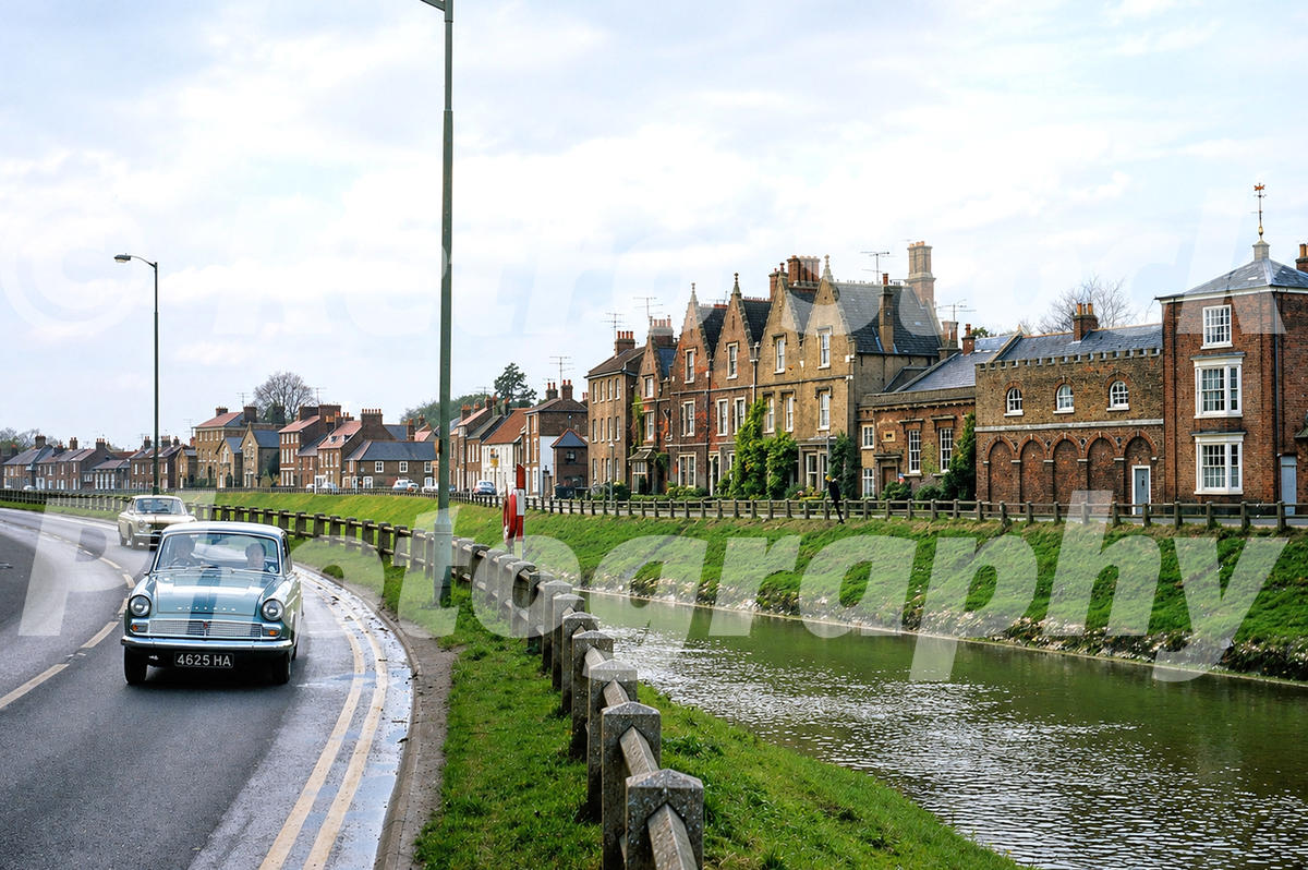 A 1970s photograph of North Brink in Wisbech, featuring a grey Hillman Minx driving alongside the River Nene and a row of historic Georgian houses in the background.